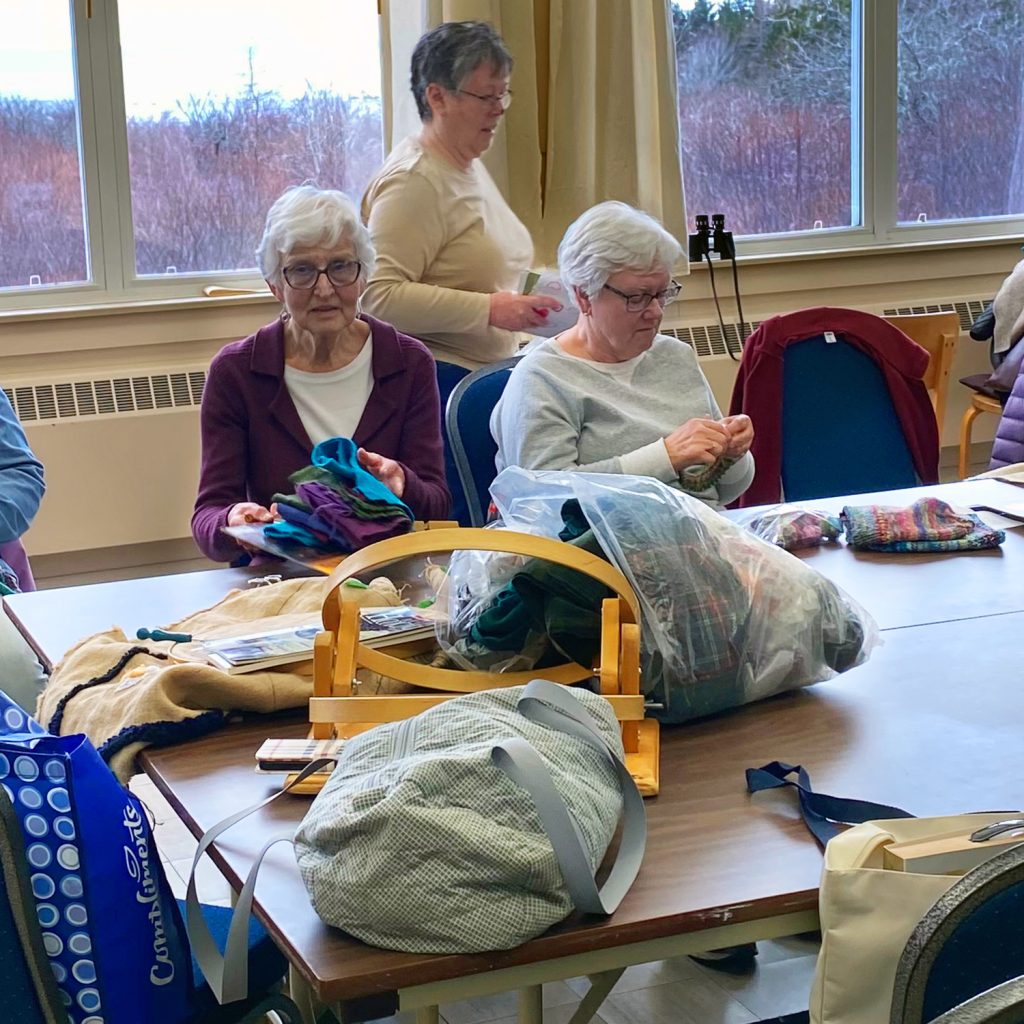 Photo of a woman at a table quilting with quilts hanging up behind her that supports the Seaside Centre Quilters. 