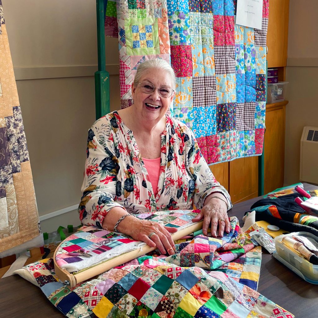 Photo of a woman at a table quilting with quilts hanging up behind her that supports the Seaside Centre Quilters. 